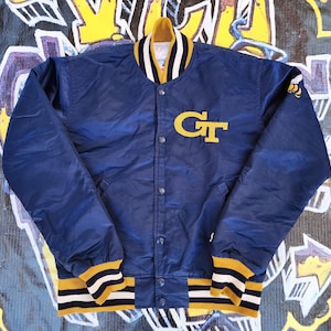 May include: A navy blue varsity jacket with a yellow and white striped collar, cuffs, and waistband. The jacket features a large yellow "GT" patch on the chest and a bee emblem on the sleeve. The jacket has a snap-button closure.