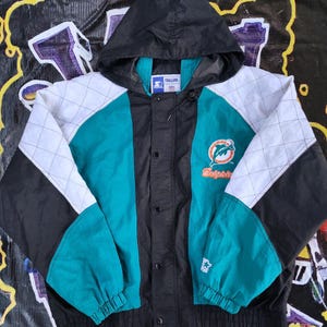 May include: A black, teal, and white hooded jacket featuring a quilted pattern on the sleeves and shoulders. The front displays the Miami Dolphins logo and text, a Starter brand logo on the cuff, and a Pro Line NFL label inside the collar.