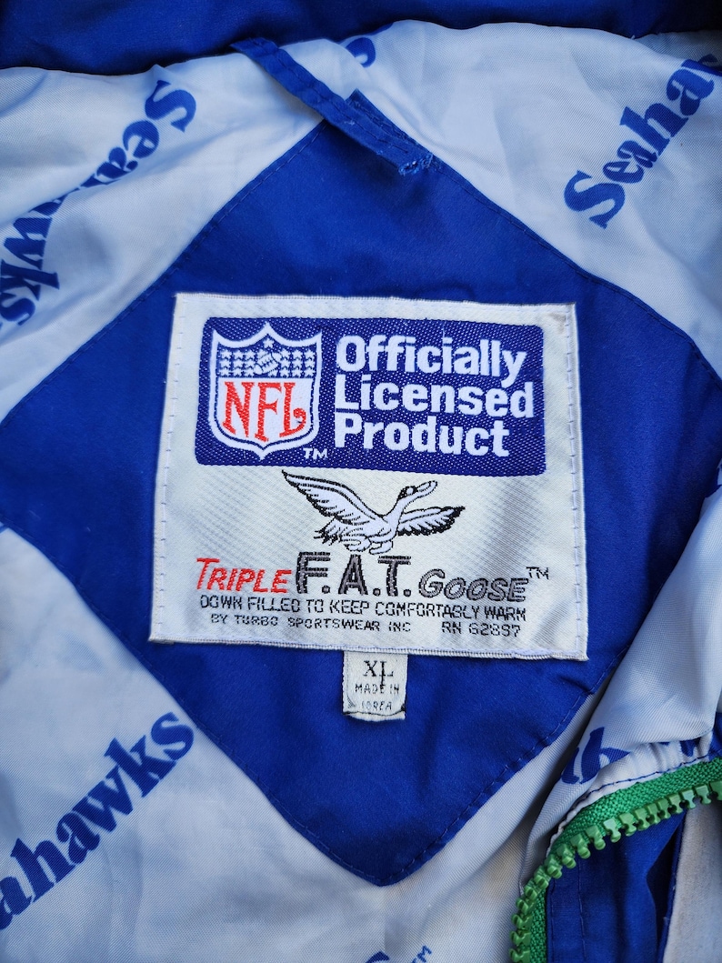 May include: Close-up of a blue and white Seahawks jacket interior. The jacket features a diamond-shaped blue panel with a white background. The NFL officially licensed product label is visible, along with the Triple F.A.T. Goose brand label.