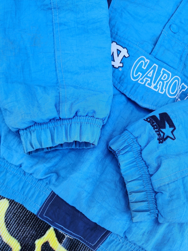 Puede incluir: Primer plano de una chaqueta azul claro con la palabra "CARO" en letras blancas. La chaqueta tiene pu&ntilde;os el&aacute;sticos y un logotipo de Starter con una estrella. La chaqueta est&aacute; hecha de un material brillante, posiblemente nailon.