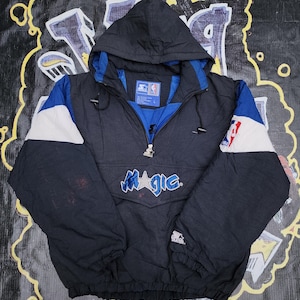 May include: A black, blue, and white hooded anorak jacket with the word "MAGIC" embroidered on the front. The jacket features a half-zip closure, a blue interior, and the NBA logo on the sleeve.