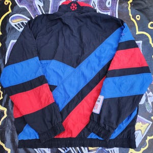Op de afbeelding: Een vintage zwarte, blauwe en rode sportjas met een hoge kraag en een voetbalontwerp. De jas heeft een colorblock-ontwerp met blauwe en rode strepen. Een kleine patch met het nummer "94" is zichtbaar.