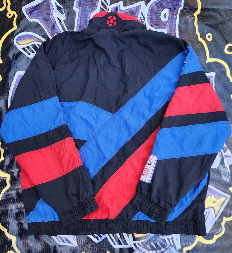 Op de afbeelding: Een vintage zwarte, blauwe en rode sportjas met een hoge kraag en een voetbalontwerp. De jas heeft een colorblock-ontwerp met blauwe en rode strepen. Een kleine patch met het nummer "94" is zichtbaar.