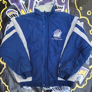 May include: A navy blue Georgetown jacket with a beige collar and accents. The jacket features a snap-button closure and a Georgetown Bulldogs logo on the chest. The sleeves have beige stripes.