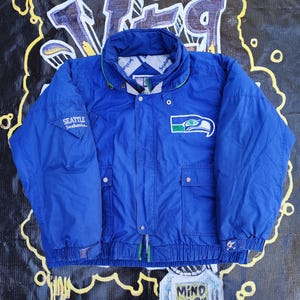 May include: A blue Seattle Seahawks jacket with a green and white logo patch on the chest. The jacket has a high collar, two front pockets, and the words "Seattle Seahawks" on the left sleeve. The jacket is a vintage style.