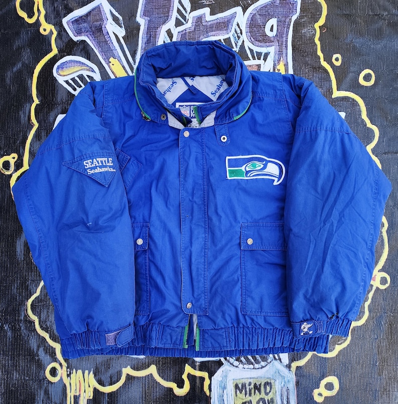 May include: A blue Seattle Seahawks jacket with a green and white logo patch on the chest. The jacket has a high collar, two front pockets, and the words "Seattle Seahawks" on the left sleeve. The jacket is a vintage style.
