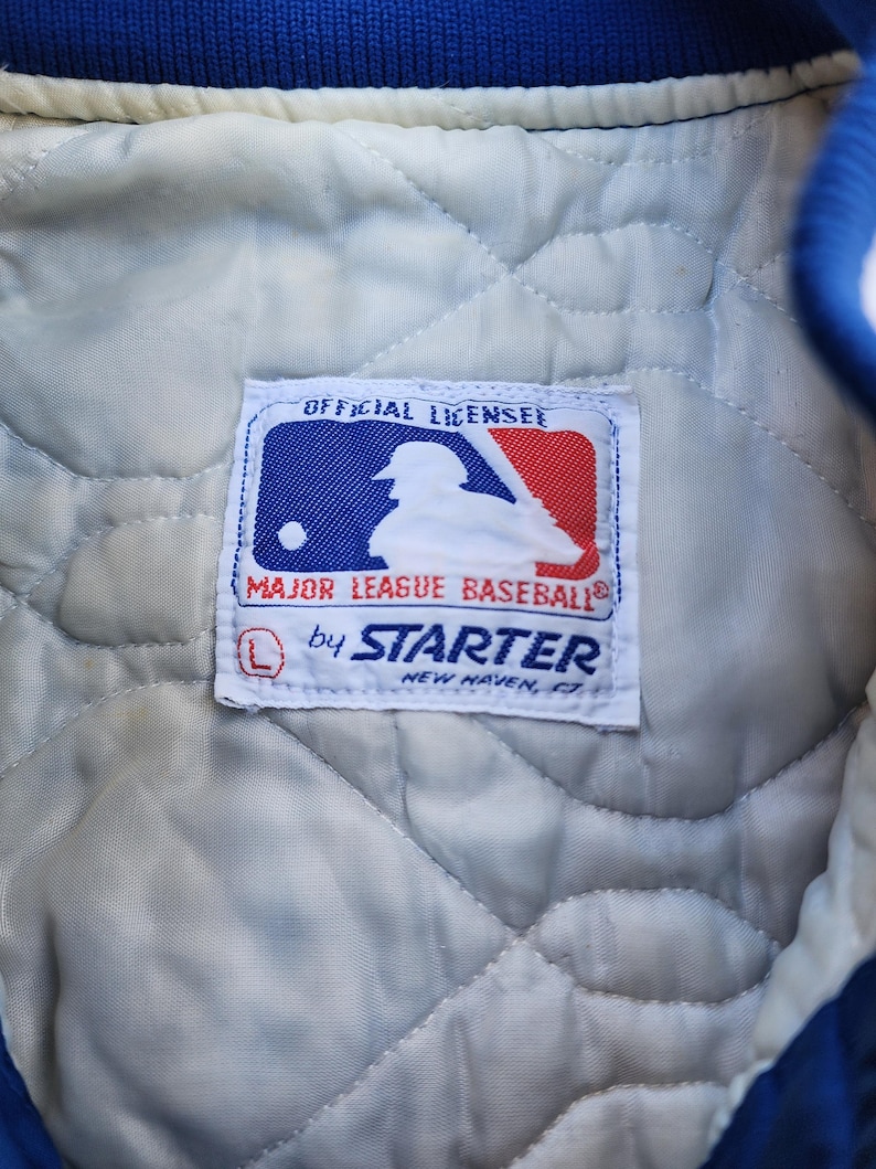 May include: A white label with blue and red trim, featuring a baseball player silhouette and the text "Official Licensee Major League Baseball by Starter New Haven, CT".