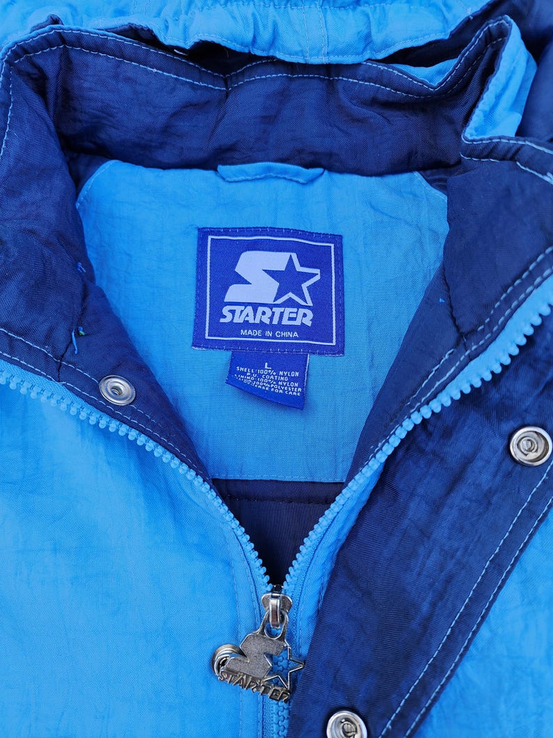 Puede incluir: Chaqueta Starter vintage en tonos azules. La chaqueta presenta una cremallera frontal, una capucha y el logotipo de Starter en la etiqueta y el tirador de la cremallera. Fabricada en nailon y poli&eacute;ster.