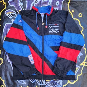 May include: A black, blue, and red vintage windbreaker jacket with a full zipper. The jacket features a color block design with the text "WorldCup USA '94" and a soccer ball graphic. The jacket has a hood and a drawstring.