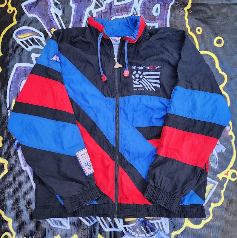 Op de afbeelding: Een vintage windjack in zwart, blauw en rood met een volledige ritssluiting. De jas heeft een colorblock-ontwerp en de tekst "WorldCup USA '94" met een voetbalafbeelding. Het heeft een capuchon en een trekkoord.