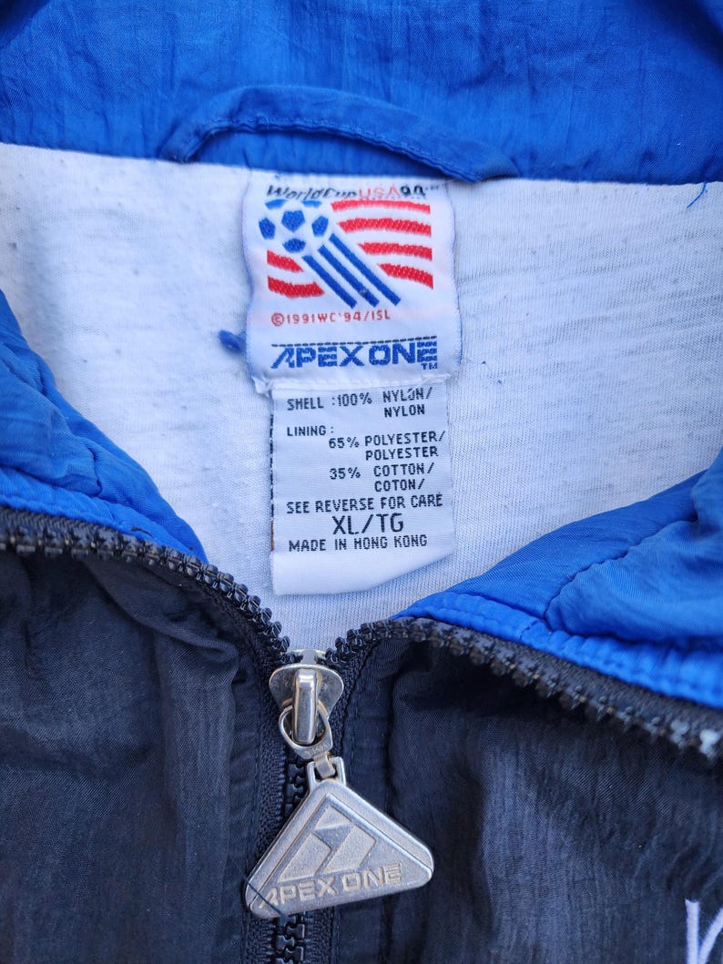 Op de afbeelding: Close-up van een vintage Apex One jassetiket. Het label toont het World Cup USA '94 logo, met een rood, wit en blauw ontwerp. De jas is zwart en blauw, met een rits en het Apex One logo op de ritstrekker.