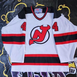 May include: White hockey jersey with black and red stripes. The jersey has a large red New Jersey Devils logo on the front with the number "4" on each sleeve.