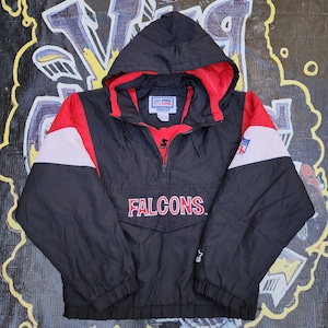 May include: A black, red, and white Atlanta Falcons pullover windbreaker jacket with a hood, a half-zip front, and long sleeves. The jacket features the word FALCONS in red lettering on the front, a Starter logo on the sleeve, and NFL branding.