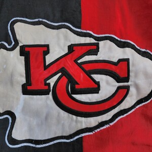 May include: A white arrowhead with a black outline and the letters "KC" in red with black outlines. The arrowhead is on a black and red background.