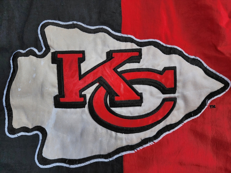 May include: A white arrowhead with a black outline and the letters "KC" in red with black outlines. The arrowhead is on a black and red background.