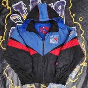 May include: A blue, red, and black New York Rangers Pro Player jacket with a hood. The jacket features the team logo on the chest and a full-length zipper. The sleeves and lower portion of the jacket are black.
