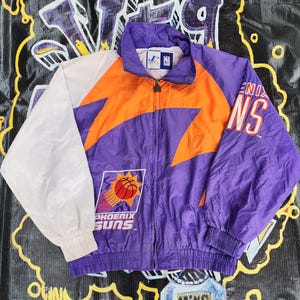 May include: A Phoenix Suns basketball team jacket in purple, orange, and white. The jacket features the team's logo on the chest and the word "Suns" below it. The sleeves have the word "Phoenix" on them.