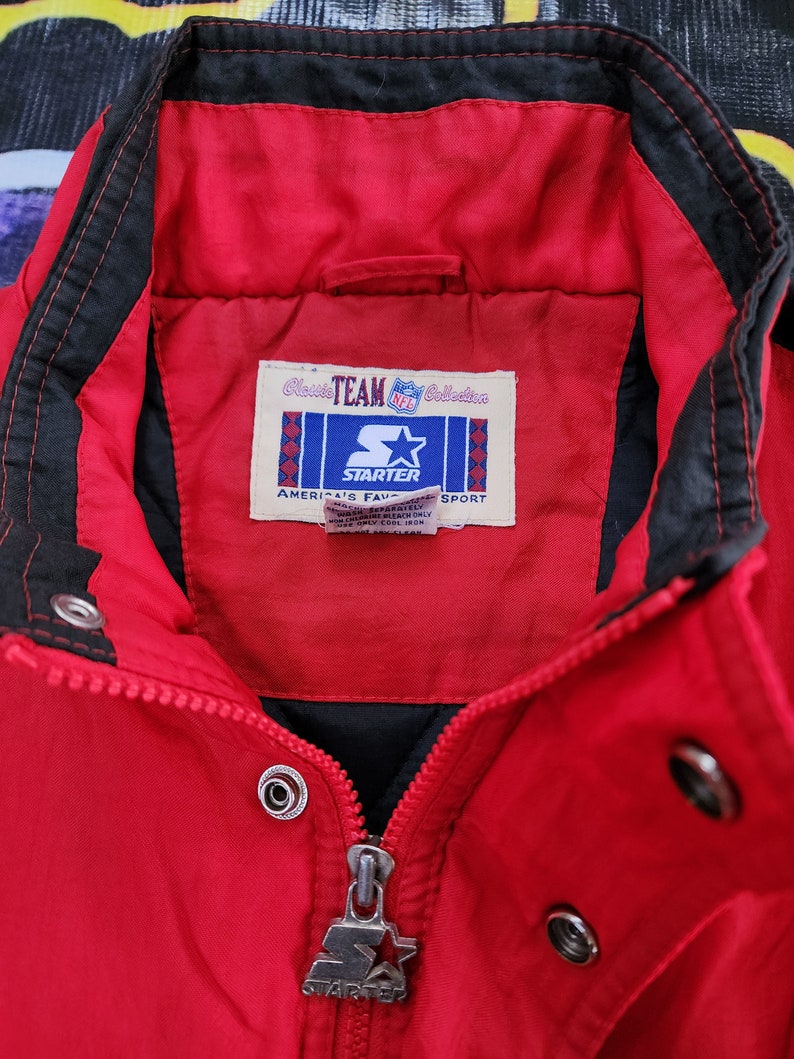May include: Red Starter jacket with a black zipper and black trim. The jacket has a label that reads "Classic Team Collection NFL Starter America's Favorite Sport".