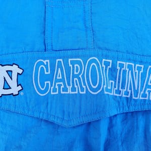 Puede incluir: Primer plano de una chaqueta azul claro con la palabra "CAROLINA" en letras blancas. La chaqueta tambi&eacute;n presenta un logotipo blanco y azul. La tela tiene un ligero brillo.