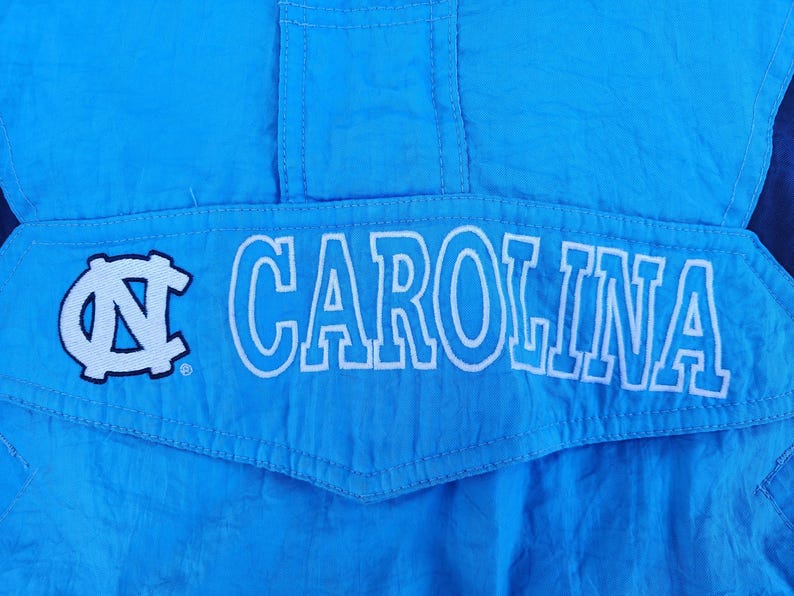 Puede incluir: Primer plano de una chaqueta azul claro con la palabra "CAROLINA" en letras blancas. La chaqueta tambi&eacute;n presenta un logotipo blanco y azul. La tela tiene un ligero brillo.