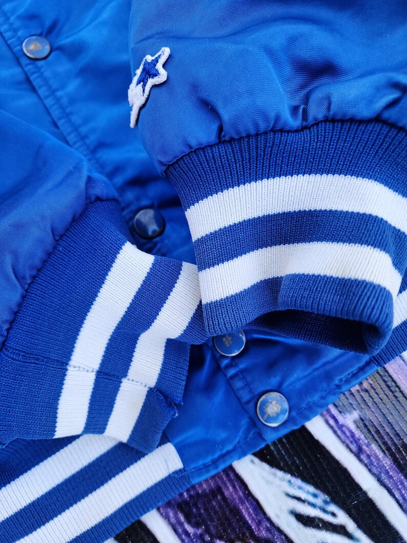 May include: Blue satin bomber jacket with white striped cuffs and a star-shaped patch on the left sleeve.