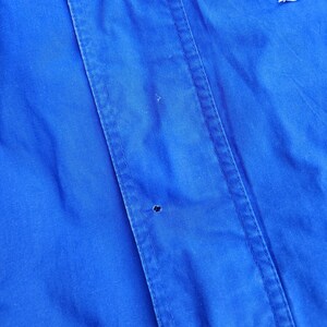 May include: Close-up of a vibrant blue jacket. The jacket features vertical seams and a small hole. A patch with white, blue, and green accents is visible at the top. The fabric appears to be a smooth, possibly water-resistant material.