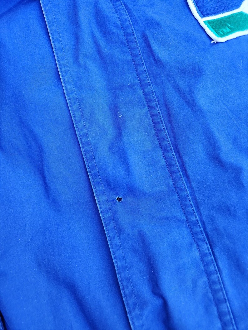 May include: Close-up of a vibrant blue jacket. The jacket features vertical seams and a small hole. A patch with white, blue, and green accents is visible at the top. The fabric appears to be a smooth, possibly water-resistant material.