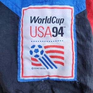 Op de afbeelding: Detail van een vintage jas met een patch met de tekst "WorldCup USA 94" en een voetbalontwerp. De patch is omlijst in blauw en rood, tegen een achtergrond van blauwe, rode en zwarte stof.