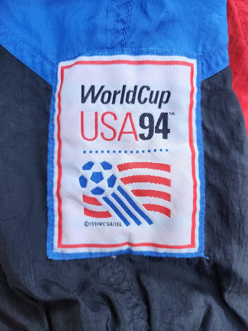 Op de afbeelding: Detail van een vintage jas met een patch met de tekst "WorldCup USA 94" en een voetbalontwerp. De patch is omlijst in blauw en rood, tegen een achtergrond van blauwe, rode en zwarte stof.