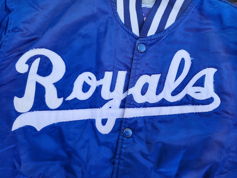 May include: A blue satin baseball jacket with a white "Royals" logo on the front.