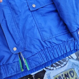 May include: A vibrant blue jacket with two front pockets and a zipper. The jacket has a ribbed hem and cuffs, and features silver buttons. The zipper has a green pull. The bottom of the jacket has the words "MIND 90".
