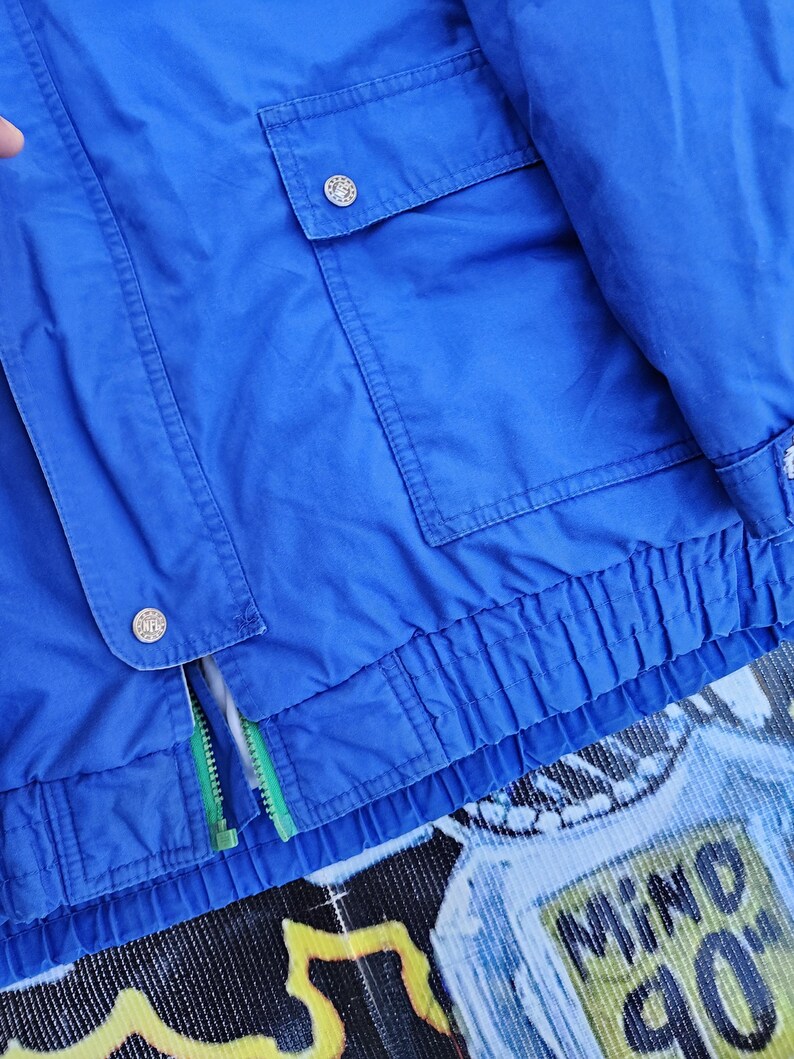 May include: A vibrant blue jacket with two front pockets and a zipper. The jacket has a ribbed hem and cuffs, and features silver buttons. The zipper has a green pull. The bottom of the jacket has the words "MIND 90".