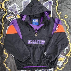 May include: Black, purple, and orange Suns anorak jacket with a hood. The jacket features the word "SUNS" across the front in white lettering. The jacket has a zipper closure and the NBA logo on the sleeve.
