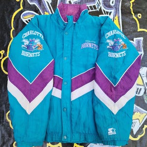 May include: Teal, purple, and white Charlotte Hornets Starter jacket. The jacket features the team logo on the sleeves and chest, with the word "Hornets" and the team name. The jacket has a button-up front and a stand-up collar. The Starter logo is on the bottom.