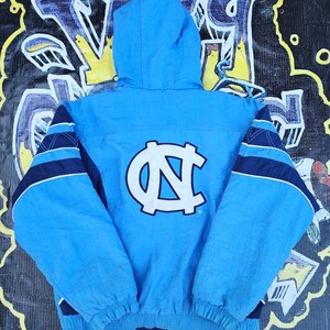 Puede incluir: Chaqueta con capucha azul claro con el logotipo de la Universidad de Carolina del Norte en la espalda. La chaqueta presenta detalles de rayas azul marino y blanco en las mangas. La chaqueta est&aacute; hecha de un material ligero y tiene un cord&oacute;n en la parte inferior.