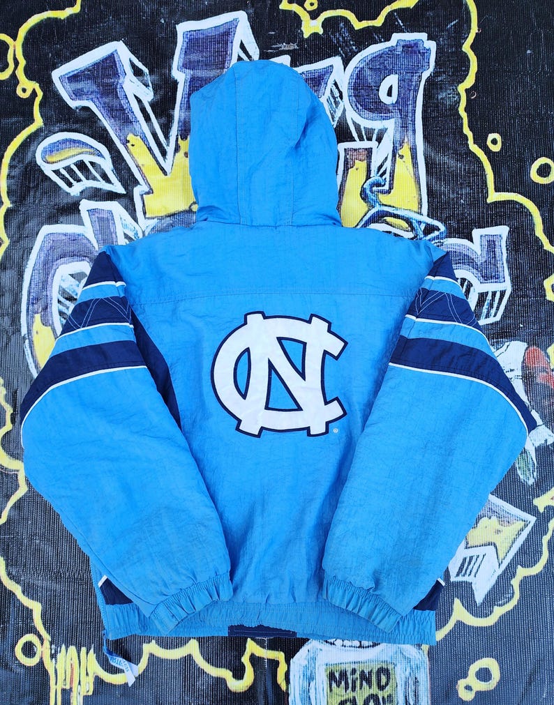 Puede incluir: Chaqueta con capucha azul claro con el logotipo de la Universidad de Carolina del Norte en la espalda. La chaqueta presenta detalles de rayas azul marino y blanco en las mangas. La chaqueta est&aacute; hecha de un material ligero y tiene un cord&oacute;n en la parte inferior.