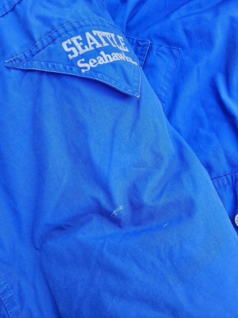 May include: Close-up of a vibrant blue jacket. A white patch on the upper left chest displays the text "SEATTLE Seahawks" in a curved font. The jacket has a simple design with a few visible seams and a button.