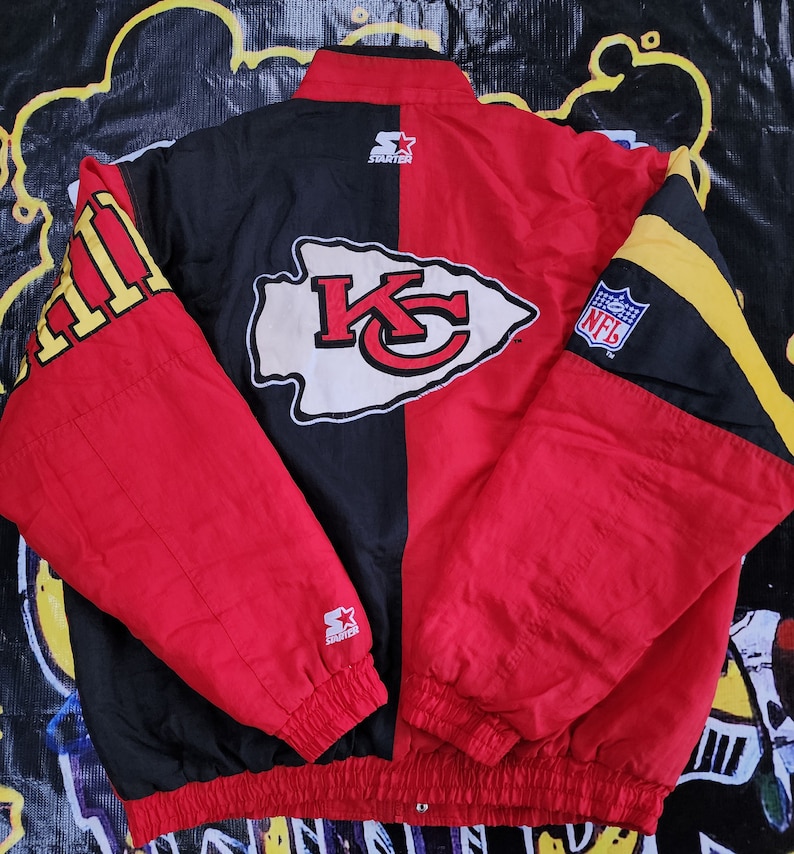 May include: A red and black Starter brand jacket with the Kansas City Chiefs logo on the back. The jacket has a yellow and black stripe on the right sleeve and the NFL logo on the left sleeve.