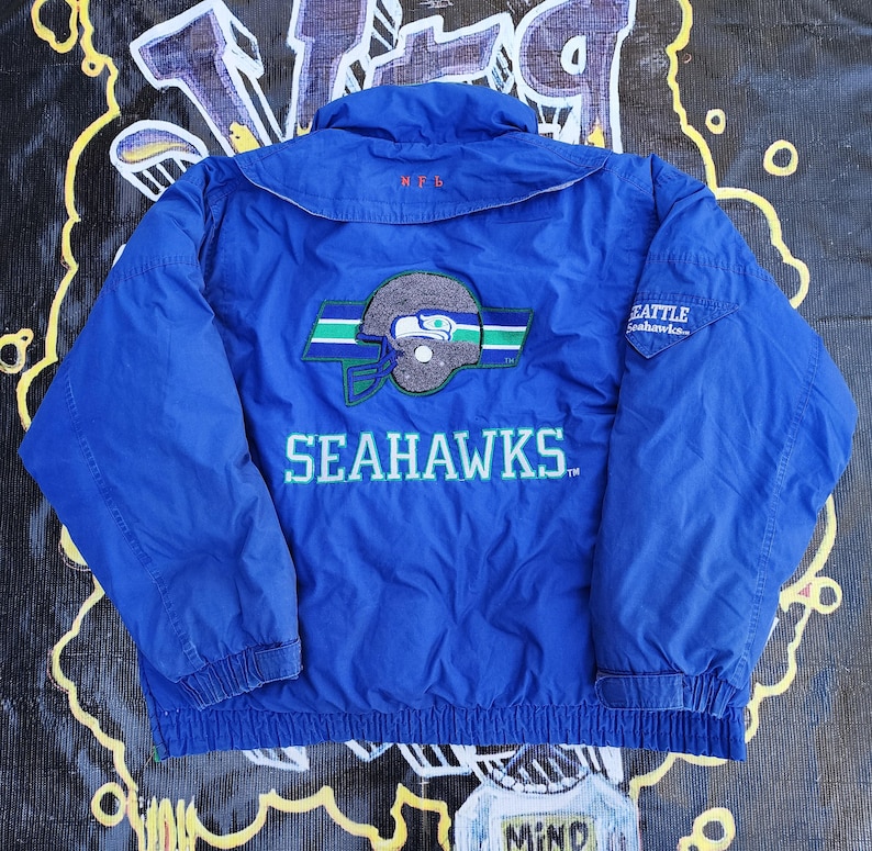 May include: A blue Seattle Seahawks jacket with the team logo on the back. The logo features a stylized helmet and the word "SEAHAWKS" in white. The jacket has a collar and two pockets.