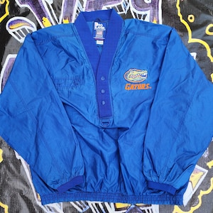 May include: A blue windbreaker jacket with a gator logo and the word "GATORS" embroidered on the front. The jacket has a snap closure and a drawstring at the bottom.