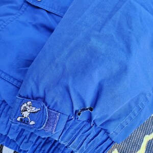 May include: Close-up of a bright blue jacket sleeve. The sleeve features a patch with a cartoon character and the letters "R.A.T." A small tear is visible near the cuff. The jacket has a buttoned pocket and a button on the shoulder.