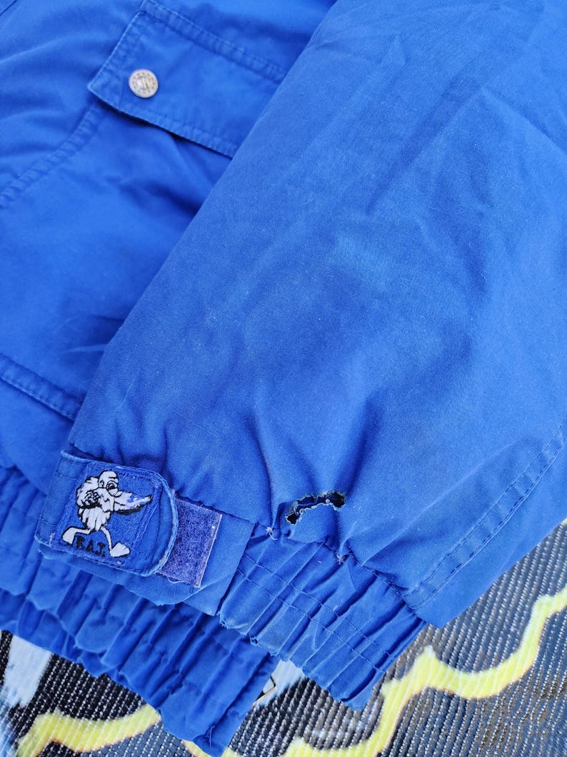 May include: Close-up of a bright blue jacket sleeve. The sleeve features a patch with a cartoon character and the letters "R.A.T." A small tear is visible near the cuff. The jacket has a buttoned pocket and a button on the shoulder.