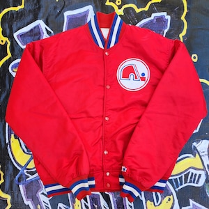 May include: A red satin baseball jacket with blue and white striped ribbing at the collar, cuffs, and waistband. The jacket has a snap closure and features a large embroidered logo on the left chest.