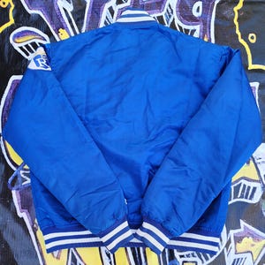 May include: A blue satin bomber jacket with white and blue striped ribbing at the collar, cuffs, and hem. The jacket has a large white and blue logo on the left sleeve.