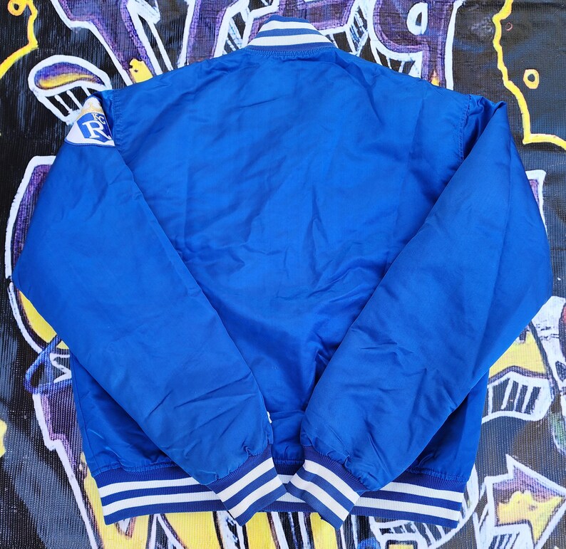 May include: A blue satin bomber jacket with white and blue striped ribbing at the collar, cuffs, and hem. The jacket has a large white and blue logo on the left sleeve.