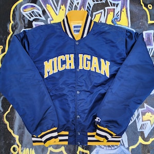 Peut inclure: Une veste de baseball en satin bleu avec des bordures rayées jaunes et blanches. La veste a une fermeture à pression et porte l'inscription "MICHIGAN" en lettres jaunes sur le devant.
