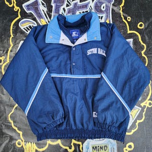 May include: A navy blue Starter pullover jacket with a light blue collar and accents. The jacket features the text "Seton Hall" and a Starter logo. The jacket has a drawstring hem and elastic cuffs.