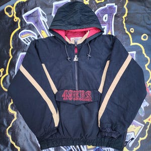 May include: Black hooded jacket with tan stripes on the sleeves and the text "49ERS" in red. The jacket has a red lining in the hood and a half-zip closure. A vintage Starter jacket.