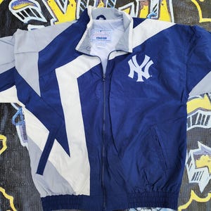 May include: A navy blue and white Starter brand jacket with the New York Yankees logo. The jacket has a full zipper, a stand-up collar, and a geometric design with gray and white accents. The jacket is made of a lightweight material.
