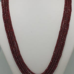 May include: A multi-strand necklace featuring five rows of small, faceted, deep red beads. The strands are gathered at the top with gold-toned metal caps and red thread accents, creating a layered, cascading design that hangs in a U-shape.